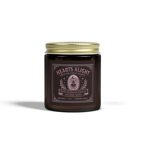 A circular, dark amber glass jar candle with a solid gold-colored metallic lid. The glass jar has a large, rectangular black label with a light-colored decorative border and ornate scrollwork. The label features the title "Hearts Alight" at the top in elegant script and lists the scent notes "Exotic Cardamom, Coconut, and Zesty Lemon" below it. A single white cotton wick with a lit flame is visible, centered within the wax. The wax itself is a rich, warm, orange-amber color, consistent with the jar's hue. T